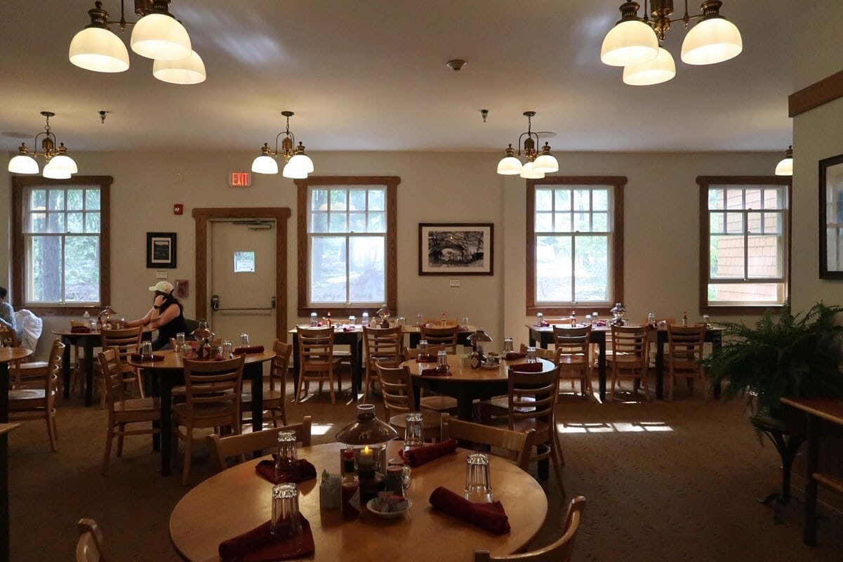 Cozy interior of the National Park Inn restaurant in Mount Rainier National Park, featuring wooden tables set with red napkins, glassware, and condiments under warm pendant lighting. Large windows let in natural light, giving diners a peaceful view of the surrounding forest.