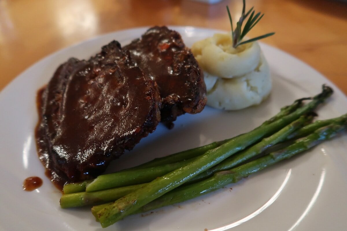 Close-up of a dinner plate featuring glazed bison meatloaf with mashed potatoes garnished with rosemary and a side of grilled asparagus. This hearty meal is one of the standout dishes often praised in National Park Inn reviews.