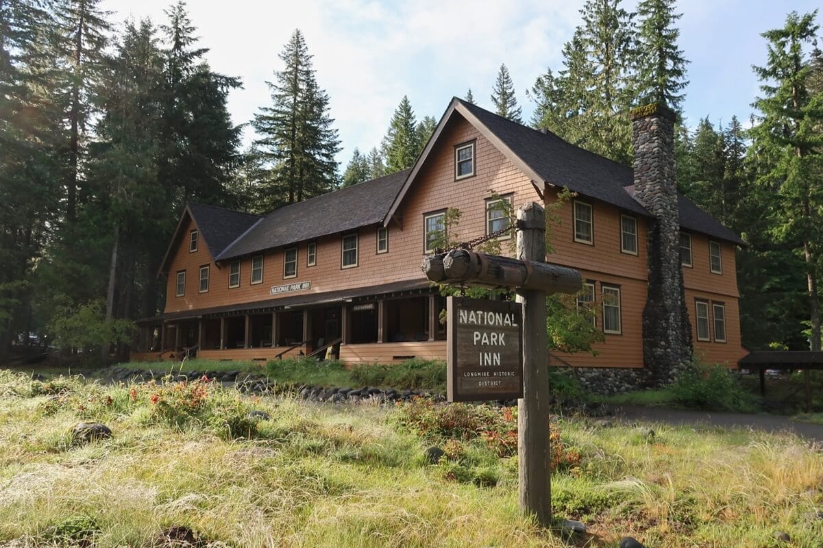 This serene setting is often highlighted in National Park Inn reviews. A rustic two-story wooden lodge in the Longmire Historic District is nestled among tall evergreens, featuring a stone chimney, a wraparound porch, and a weathered wooden sign out front.