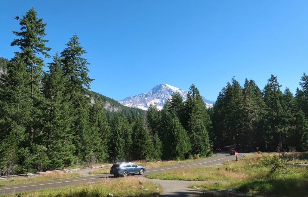 View of Mount Rainier from the National Park Inn porch shows the snow-capped peak rising above a dense evergreen forest under a bright blue sky. A winding road and stop sign frame the foreground with cars approaching.
