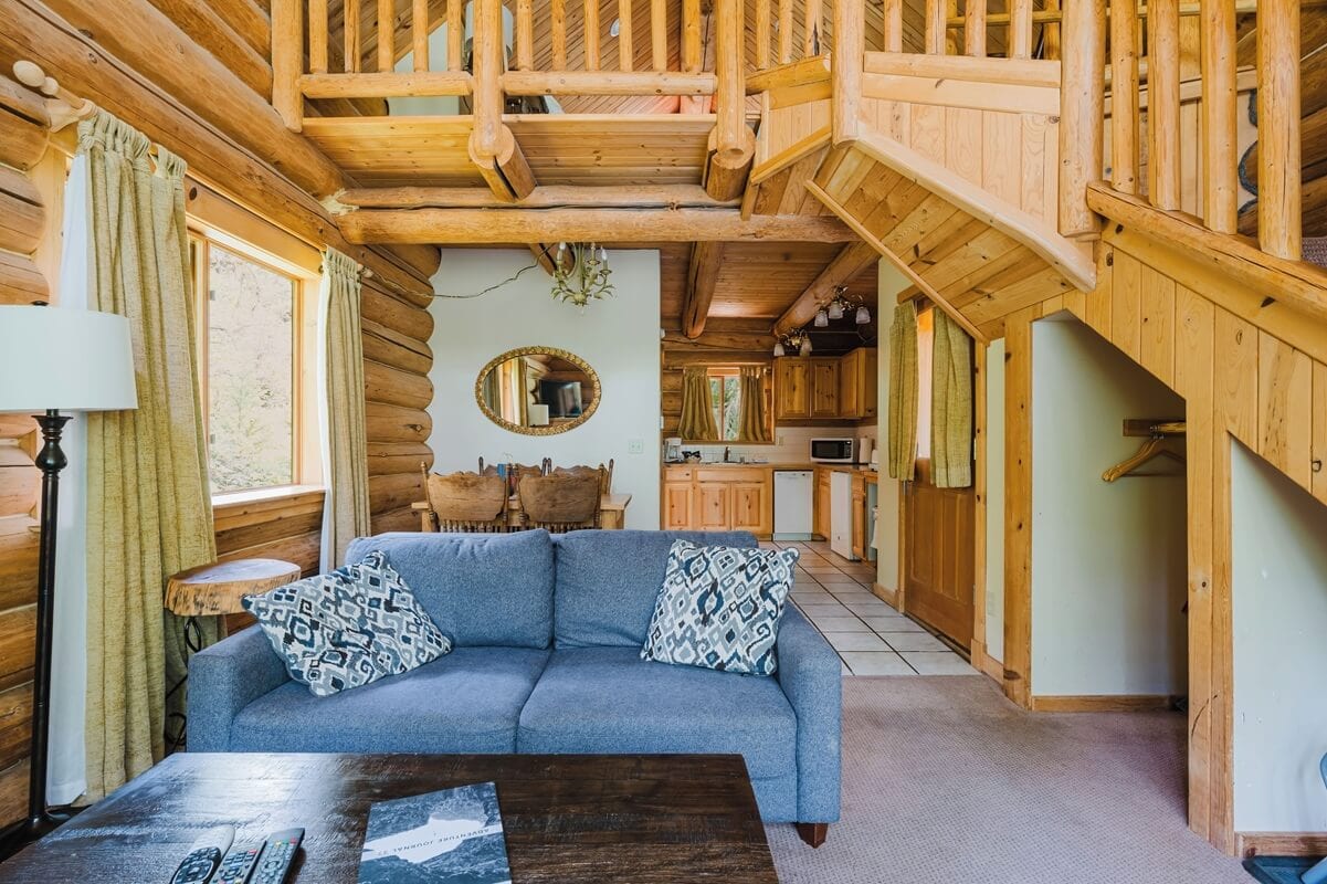 Cabins near Mount Rainier, featuring a lofted wooden staircase, rustic furniture, and a small kitchen and dining area in a log cabin interior. Natural wood tones and large windows create a warm, inviting mountain retreat.