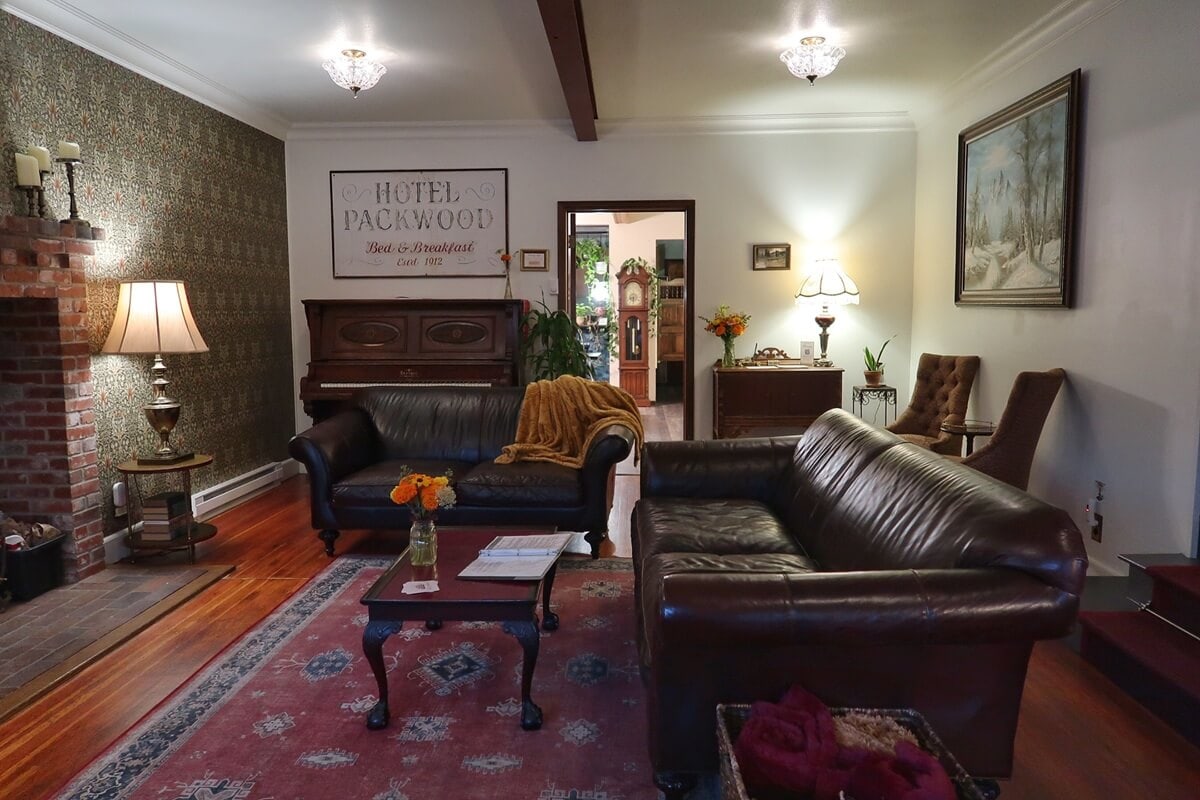 Vintage-style lounge at Historic Hotel Packwood, offering charming lodging near Mount Rainier. Warm lighting and floral accents enhance the inviting atmosphere, with leather sofas, a red area rug, antique decor, and a classic piano.