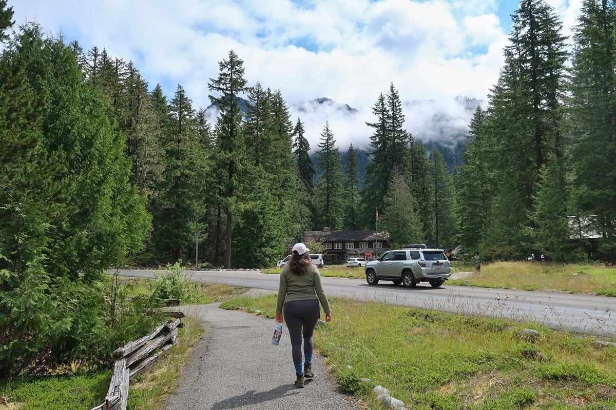 A woman walks along a paved path toward the Trail of the Shadows, surrounded by lush evergreens and mist-covered mountains. The Longmire Wilderness Information Center appears ahead, partially hidden among the trees, with a few passing cars on the nearby road.