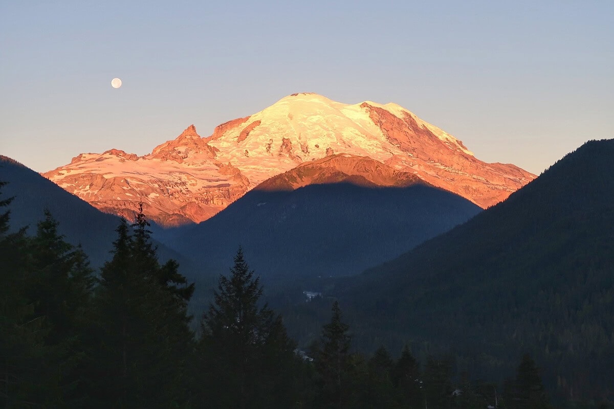 Mount Rainier at sunrise glows in warm golden and pink tones as the early light hits its snow-covered peak. Dark forested ridges frame the mountain, with a full moon still visible in the soft morning sky.