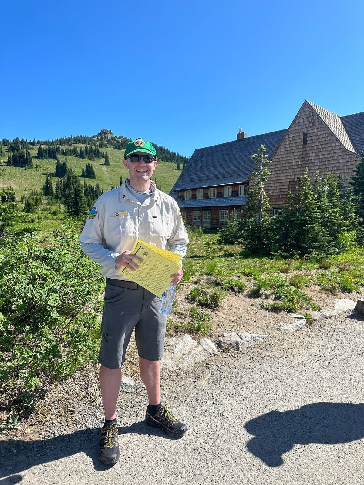 A smiling volunteer ranger helps guests learn how to visit Mount Rainier. He stands on a path near the Sunrise Day Lodge, holding trail information and a water bottle, surrounded by alpine meadows and evergreen trees under a clear blue sky.
