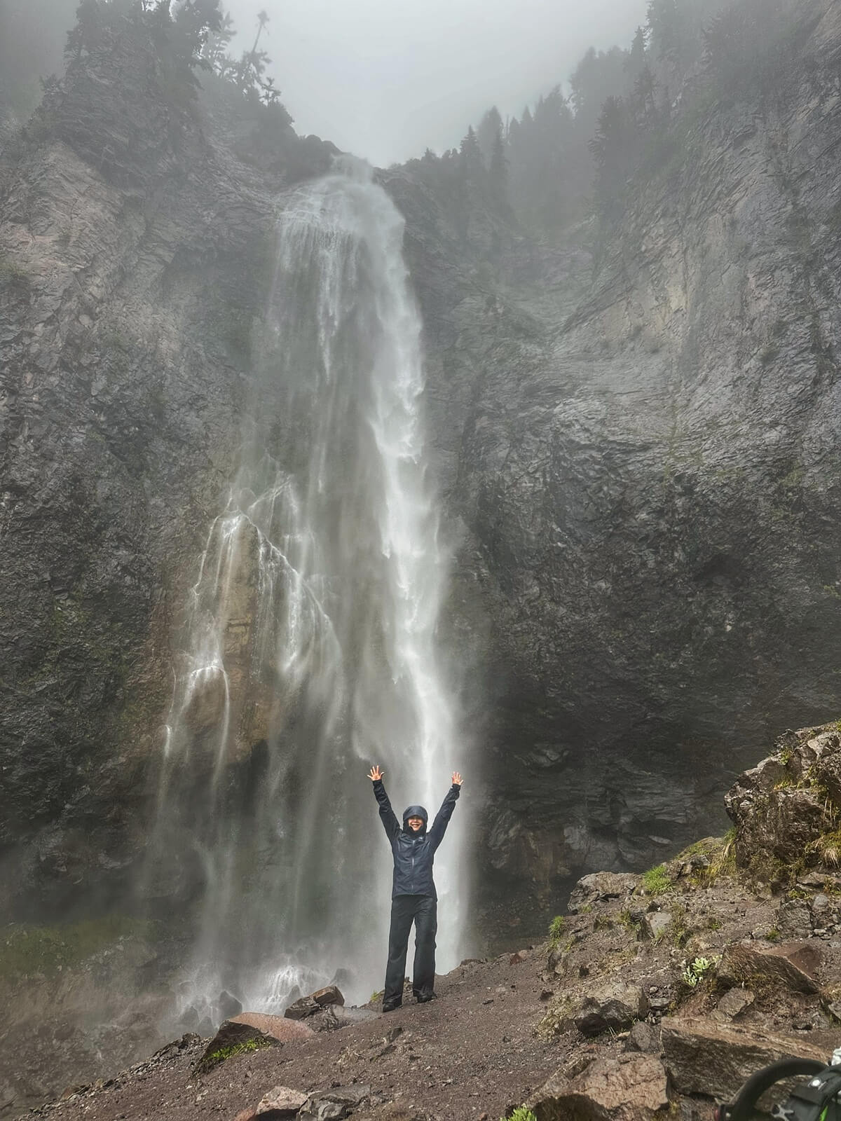 Visiting Mount Rainier on a rainy day offers dramatic waterfall views like this one, where a hiker in rain gear stands triumphantly at the base of Comet Falls. The tall cascade is surrounded by wet rock walls and a misty atmosphere.