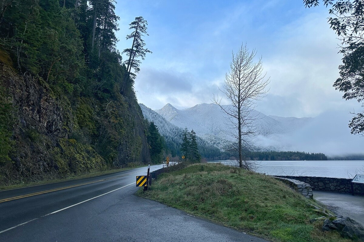 A winding road hugs a mossy cliffside in Olympic National Park in March, with a misty lake and snow-dusted mountains rising in the distance. Low clouds cling to the forested slopes as early spring blends winter's chill with signs of new growth.