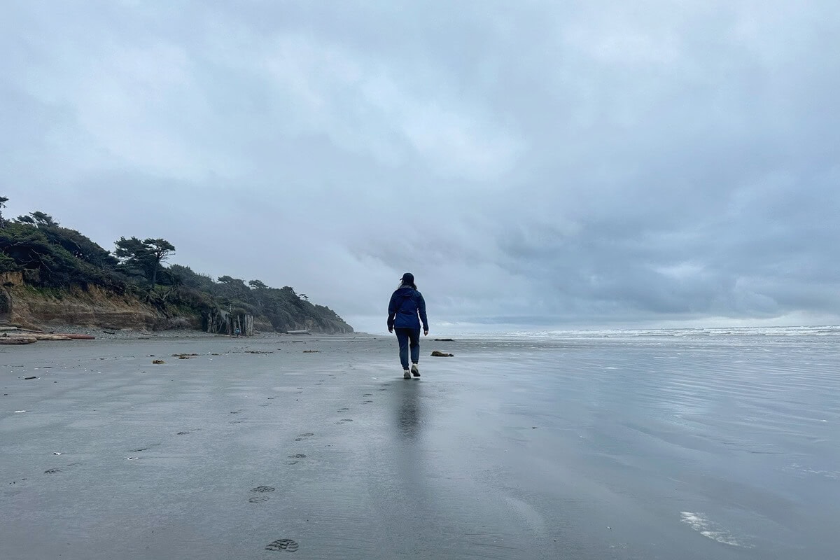 A person walks along a beach in Olympic National Park in spring, with wet sand reflecting the overcast sky. Wind-swept trees line the cliffs on the left, while waves roll in from a silvery-gray ocean under dramatic clouds.