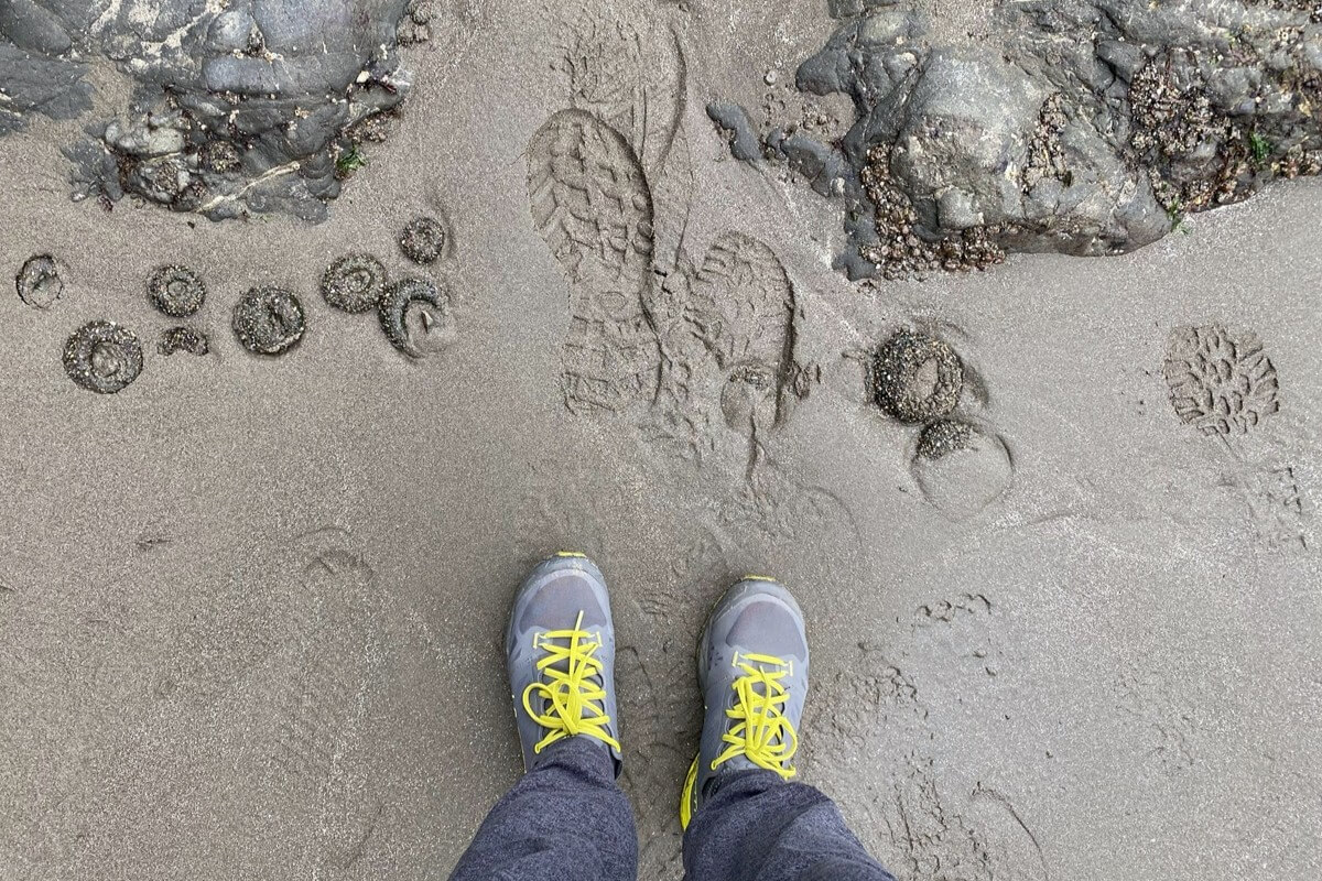 A person wearing gray hiking shoes with bright yellow laces stands on wet sand near rocky tidepools, beside boot prints and exposed sea anemones. The scene captures a close-up moment of beach exploration.