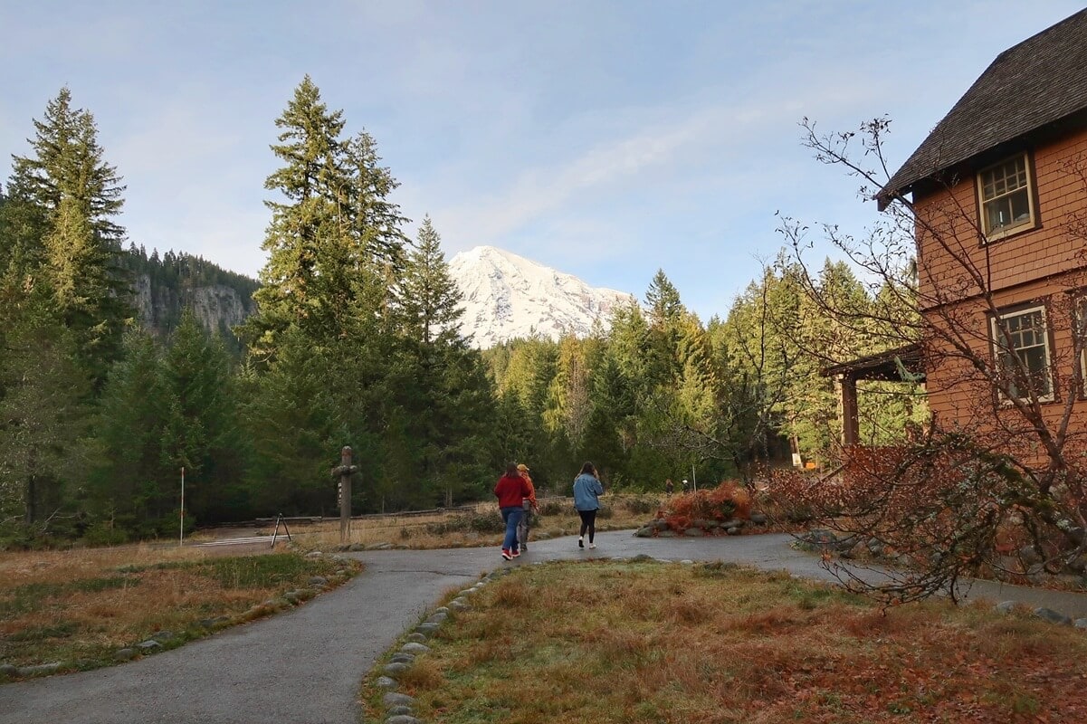 People walking along a paved path in Longmire at Mount Rainier, surrounded by tall evergreen trees with a snow covered mountain rising in the background. A rustic lodge building sits to the right, adding warmth and scale to the peaceful forest setting.