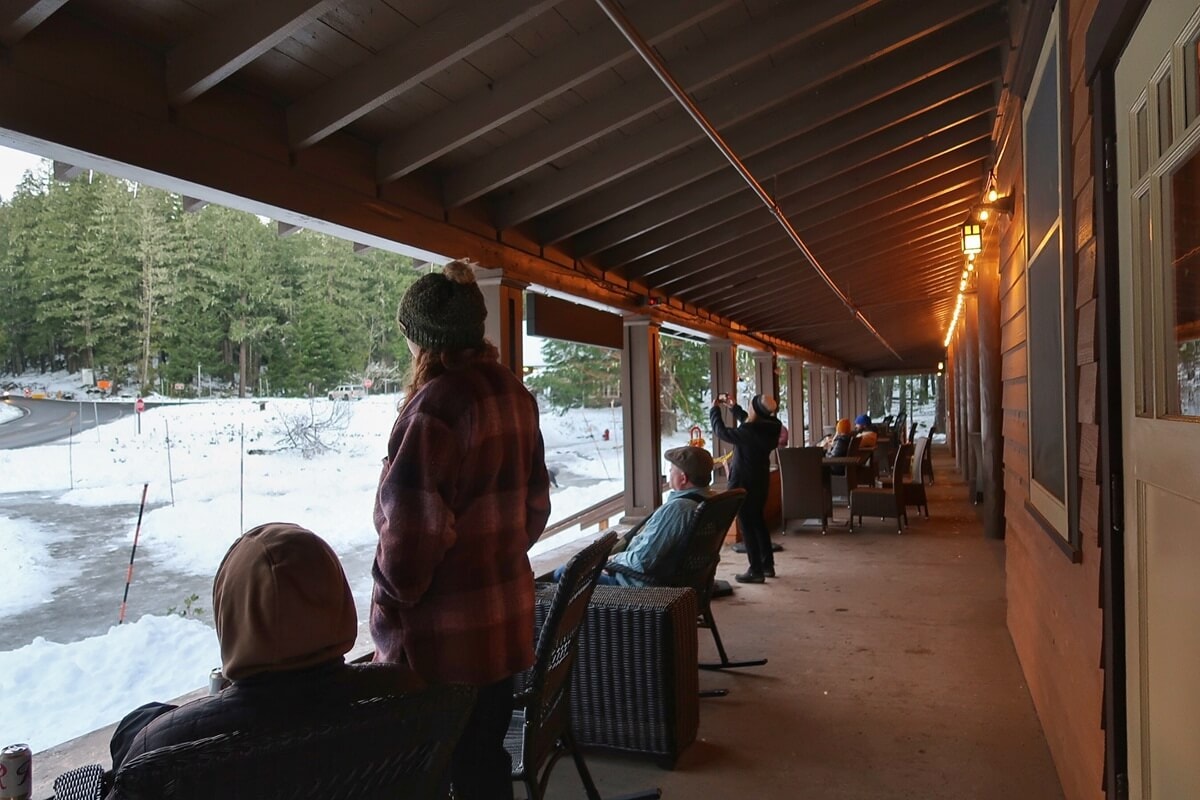 People bundled in winter jackets and knit hats relax on the covered wooden porch at National Park Inn. Warm porch lights glow above wicker chairs as visitors watch the snow and forested road, capturing a cozy winter scene at a historic park lodge.