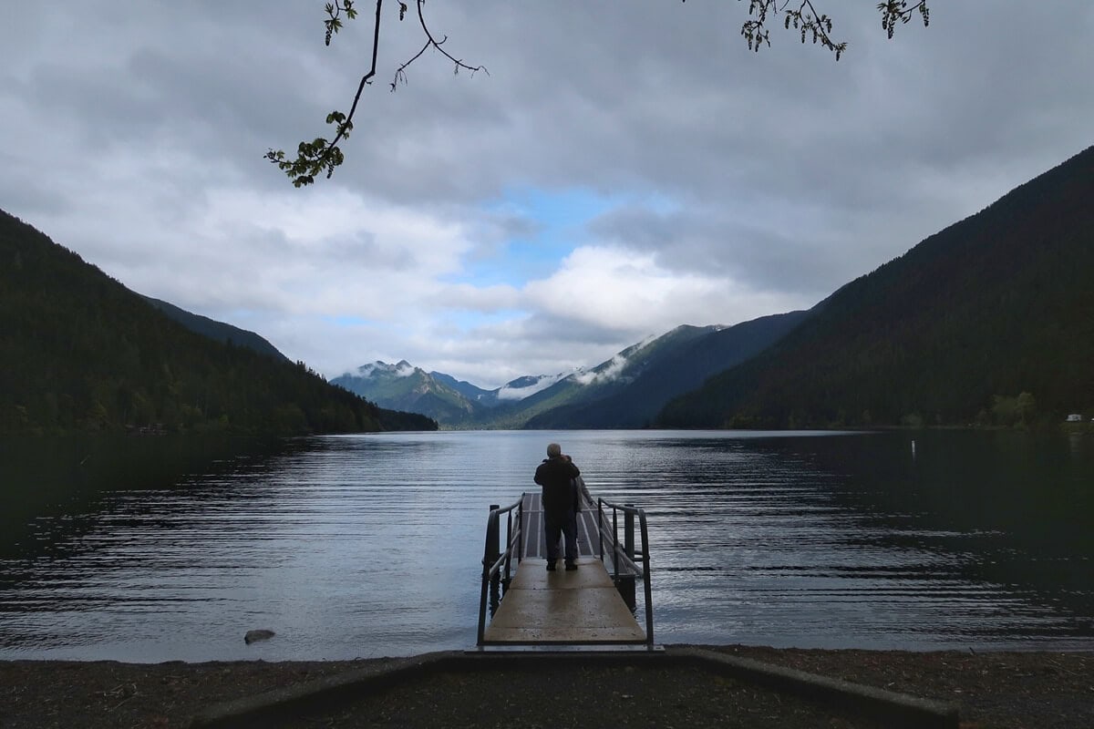 Couple standing at the end of a small dock at Fairholme boat launch looking out over a calm lake, with forested mountains and low clouds in the distance under an overcast sky.