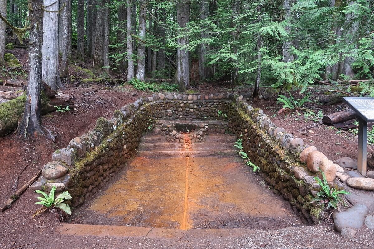 Iron Mike Springs sits beneath tall evergreen trees, with moss covered rock walls enclosing shallow mineral stained water. The orange tinted flow running through the center highlights the iron rich minerals that give the springs its distinctive color and historic significance.