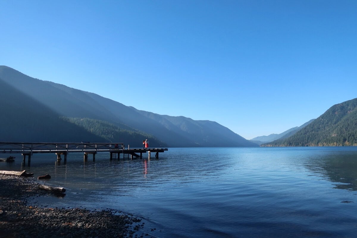 Wooden dock extending into Lake Crescent with two people standing at the end, surrounded by forested mountains under a clear sky. Sunlight casts soft rays across the lake and hills, creating a peaceful scenic view.