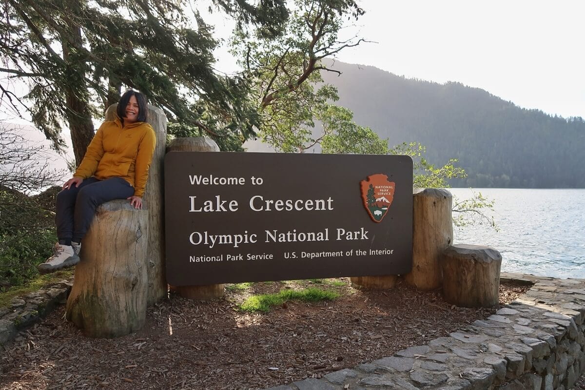 Person sitting beside a sign that says “Welcome to Lake Crescent Olympic National Park National Park Service U.S. Department of the Interior,” with a calm lake surrounded by forested mountains in the background.