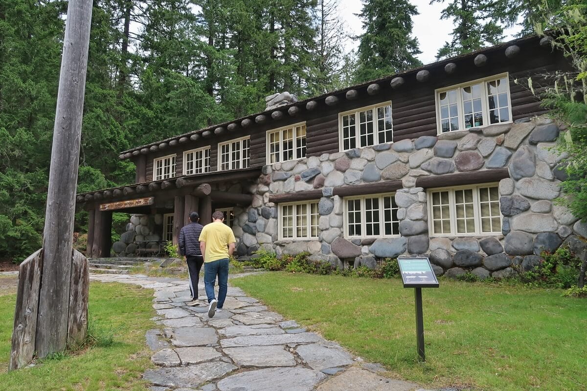 Two visitors walk along a stone pathway toward the Longmire Administration Building, a historic structure within the Longmire area of Mount Rainier National Park built with large river stones and dark wooden beams.