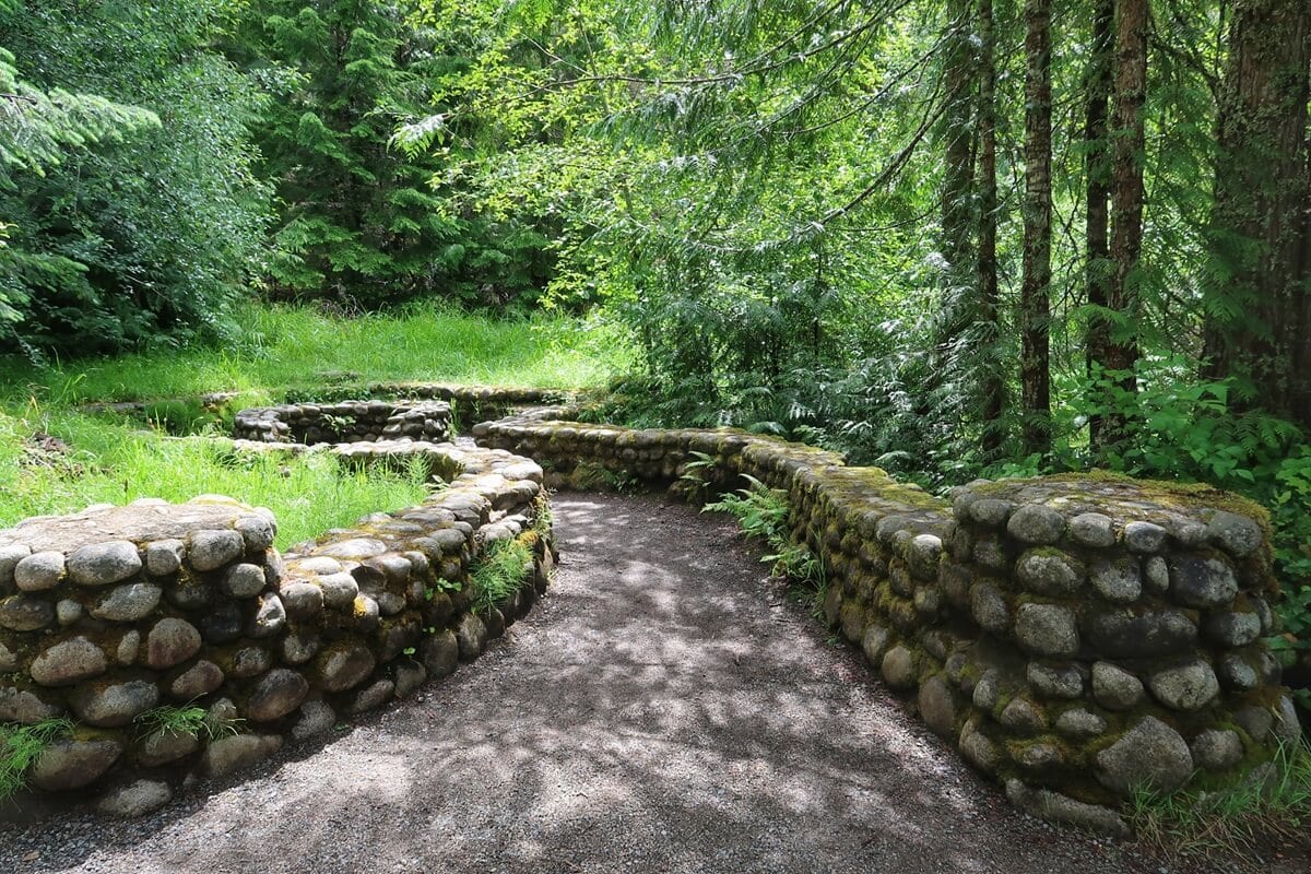 A curved stone walkway winds through a lush forest, bordered by low rock walls covered in moss and dappled sunlight. These remnants of Longmire Medical Springs Resort highlight the historic foundations nestled among towering evergreens and dense green undergrowth.