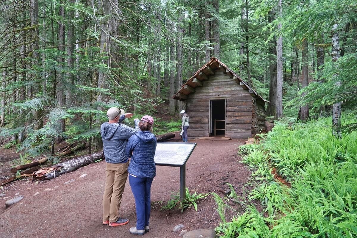 Two visitors stand near an interpretive sign in front of a rustic Longmire cabin, surrounded by tall evergreen trees and lush ferns. One person points toward the historic wooden structure while another visitor walks toward the open doorway.
