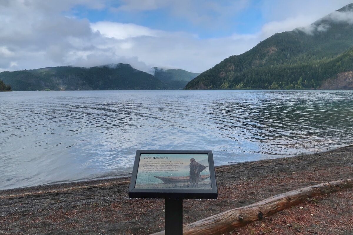 Informational sign along Moments in Time Trail overlooking a calm lake with forested mountains and low clouds in the distance, set on a pebbled shoreline with a fallen log nearby.