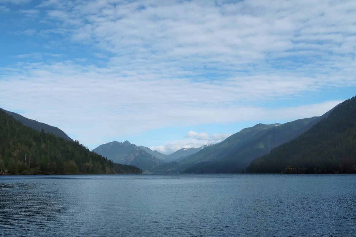 Wide Lake Crescent view from Fairholme boat launch with calm blue water stretching toward layered forested mountains under a partly cloudy sky, creating a peaceful scenic landscape.