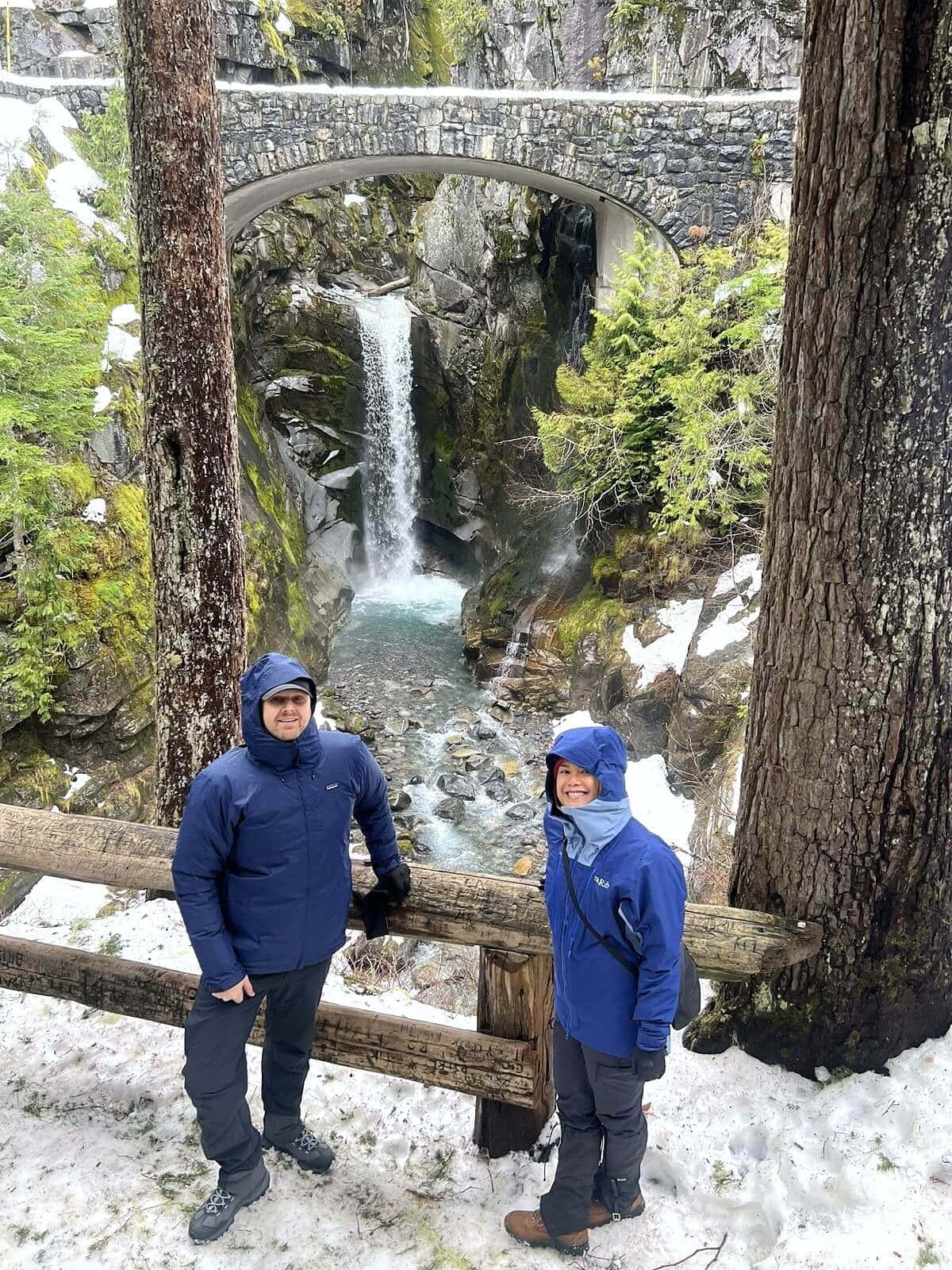 Two people in winter jackets stand by a wooden railing overlooking Christine Falls in winter as water cascades beneath a stone bridge into a rocky pool. Snow patches cling to the ground and surrounding forest, with tall trees framing the scenic waterfall viewpoint.
