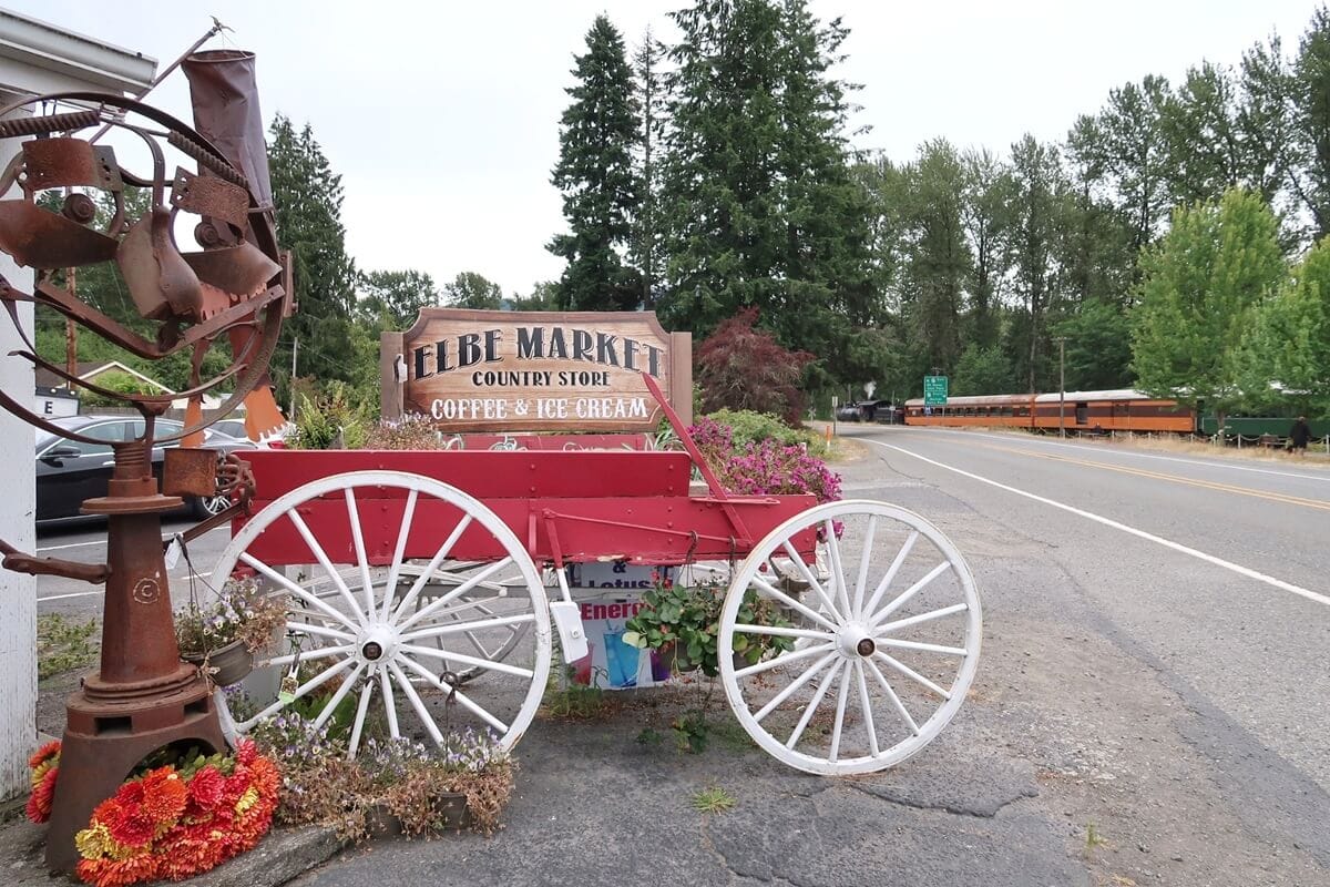 A rustic roadside display features a red wagon with white wheels filled with flowers and a wooden sign that reads “Elbe Market Country Store Coffee & Ice Cream.” A road runs alongside the small town setting with a train visible in the background.