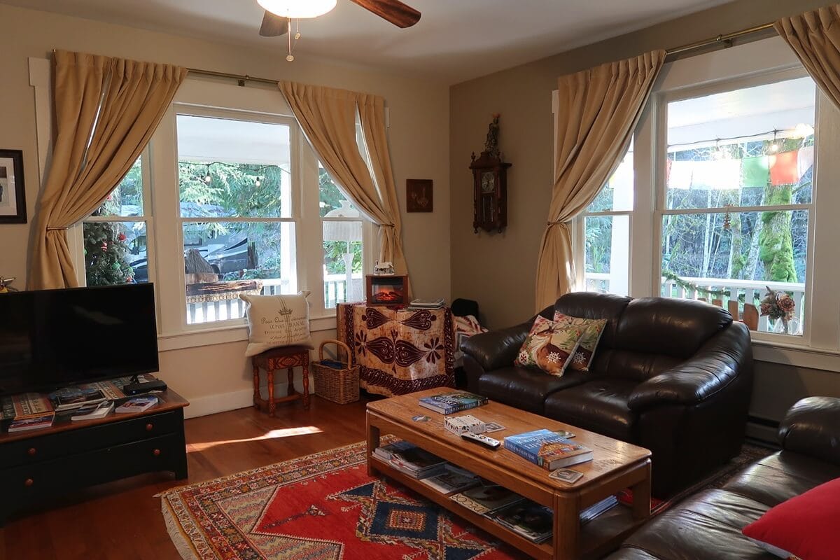 Cozy living room inside Mountain Meadows Inn in Ashford with leather seating, a wooden coffee table and warm natural light coming through large curtained windows. Decorative pillows books and a patterned rug create a comfortable inviting space with views of greenery outside.