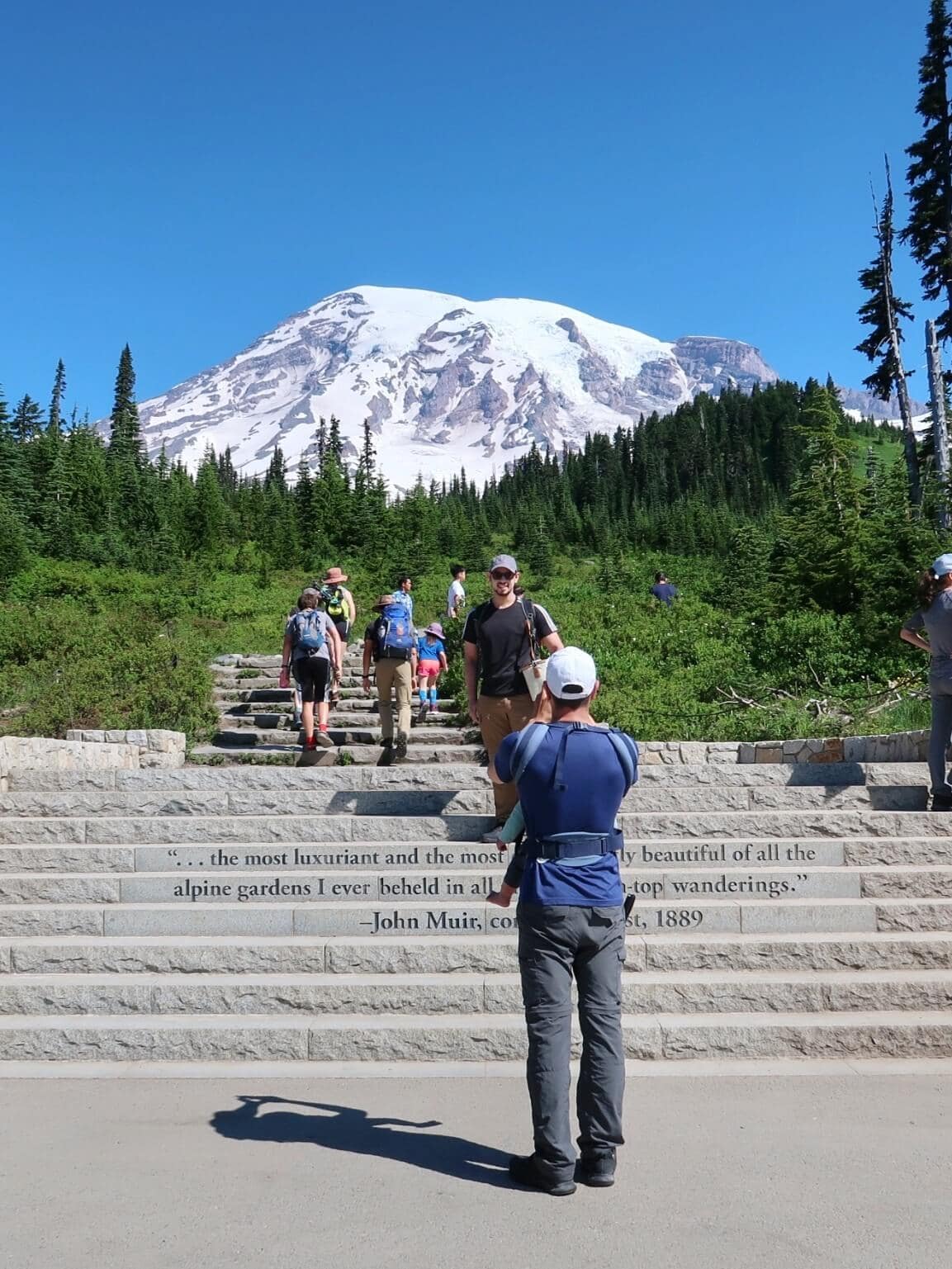 Visitors walk up the Muir Steps at Mount Rainier surrounded by a green alpine landscape. A man stands at the bottom holding a child while another person takes a photo.