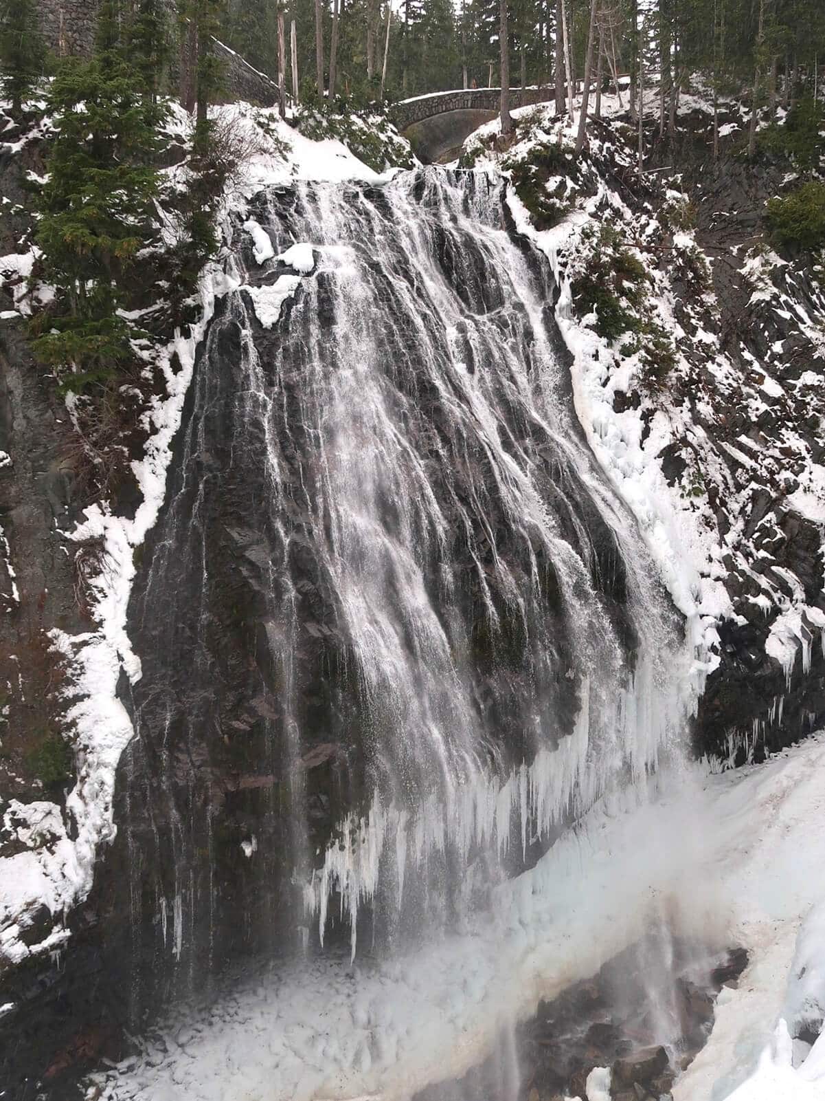 Narada Falls in winter cascades down a wide dark rock face with sections of ice forming along the edges and base of the waterfall. Snow covers the surrounding cliffs and forest while a stone bridge sits above the falls, framing the dramatic frozen landscape.