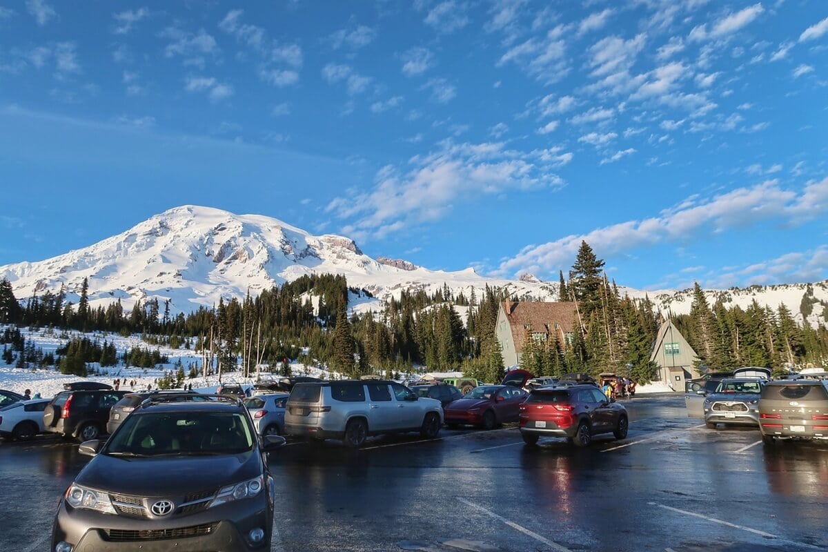 Paradise parking lot shows wet pavement and cars that drove Mount Rainier roads in winter surrounded by snow and evergreen trees. In the background a snow covered mountain rises under a blue sky while visitors gather near a rustic building at the edge of the lot.