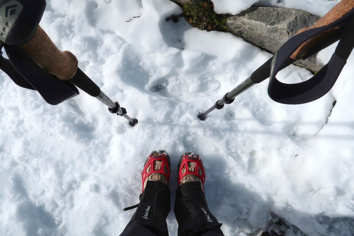 Close up view of gear for winter hikes at Mount Rainier showing microspikes strapped to boots standing in packed snow with trekking poles planted on either side. The textured snow surface highlights the practical equipment needed for stability and traction in winter conditions.