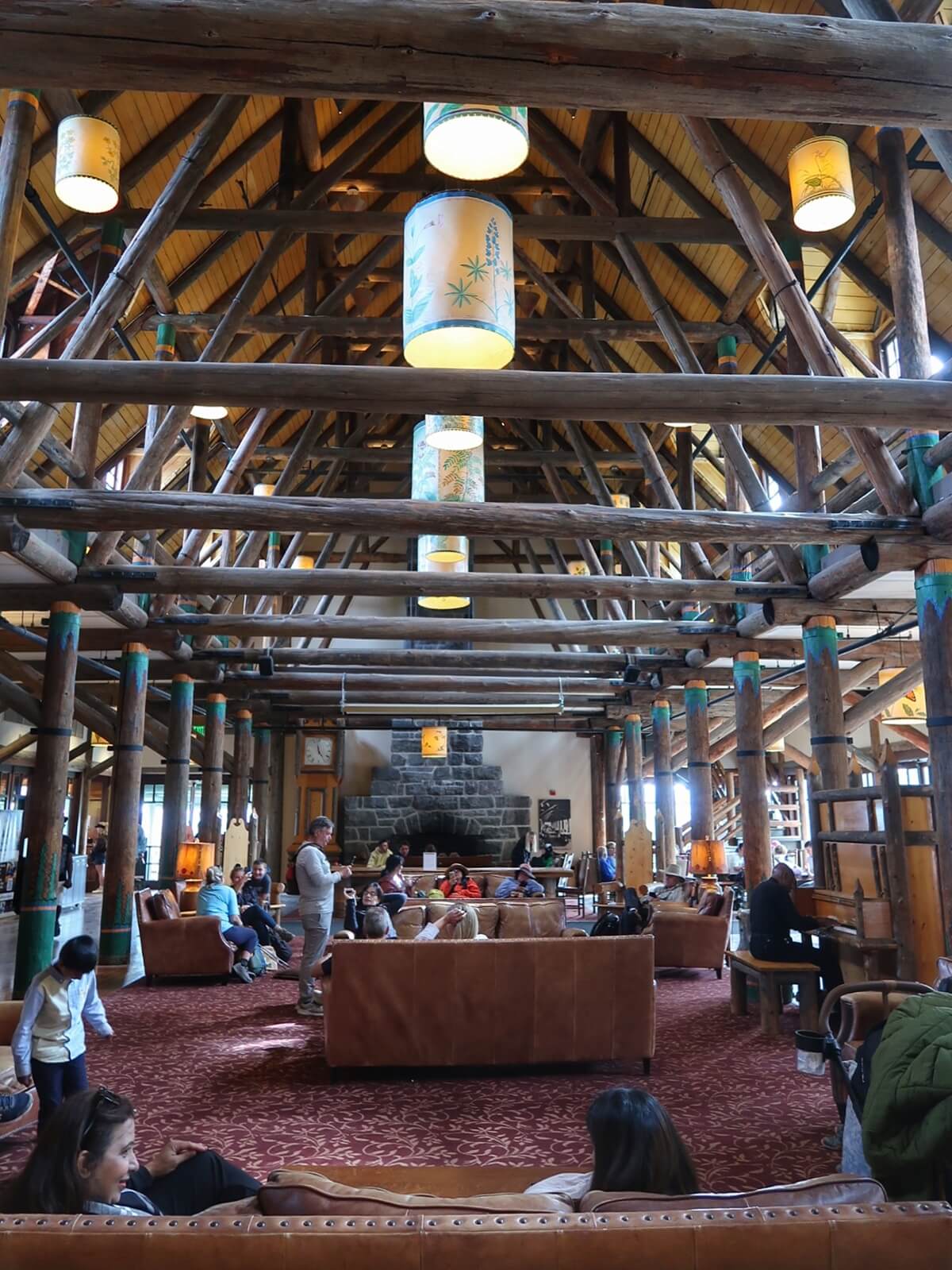 Spacious interior of Paradise Inn with exposed wooden beams, hanging lantern lights, and a large stone fireplace at the center. Visitors sit on leather couches and chairs relaxing, while others walk through the warm and rustic gathering space.