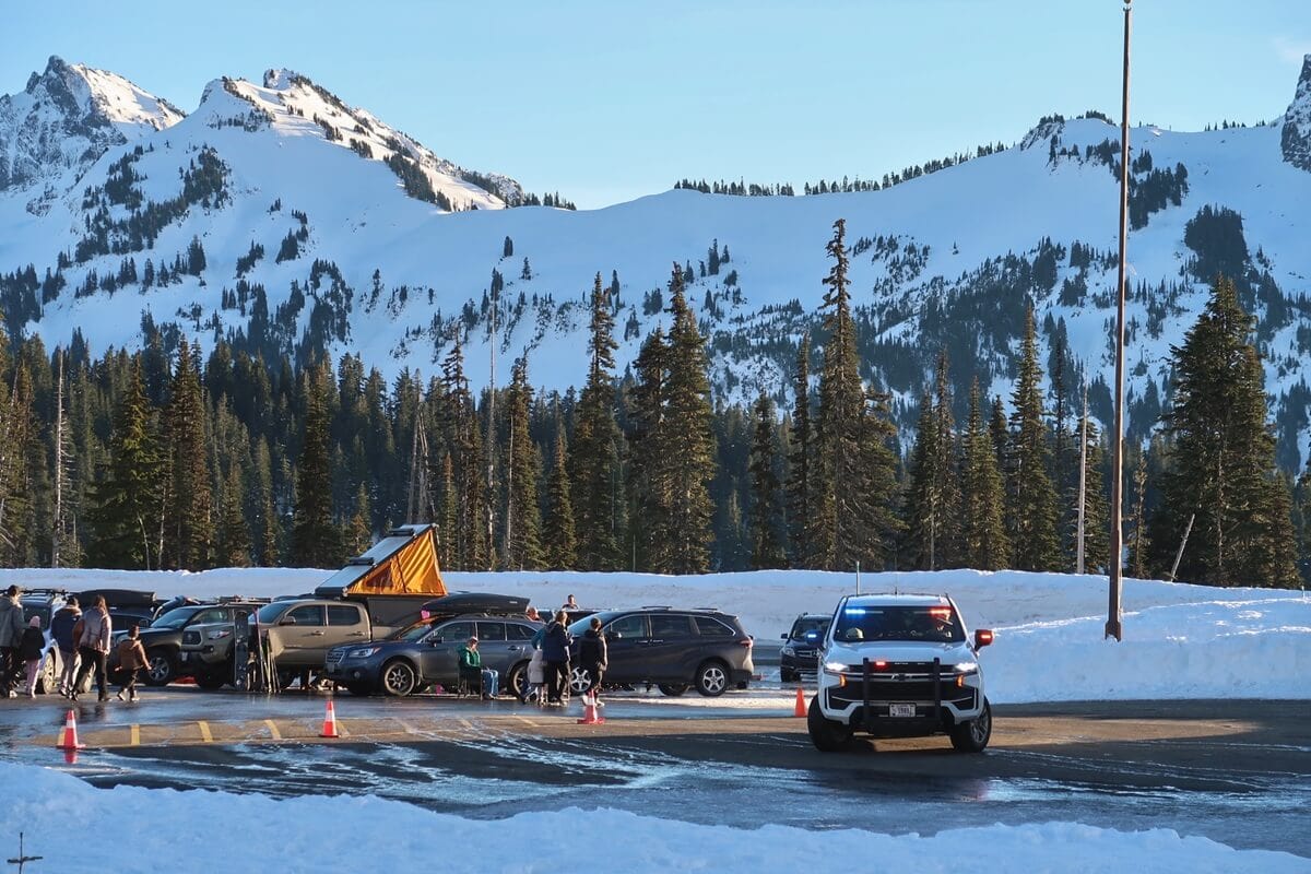 A snowy parking area with cars and people gathered near the edge of a plowed lot while a ranger vehicle with flashing lights sits in the foreground. Behind them a line of evergreen trees leads up to steep snow covered mountain peaks under a clear sky.