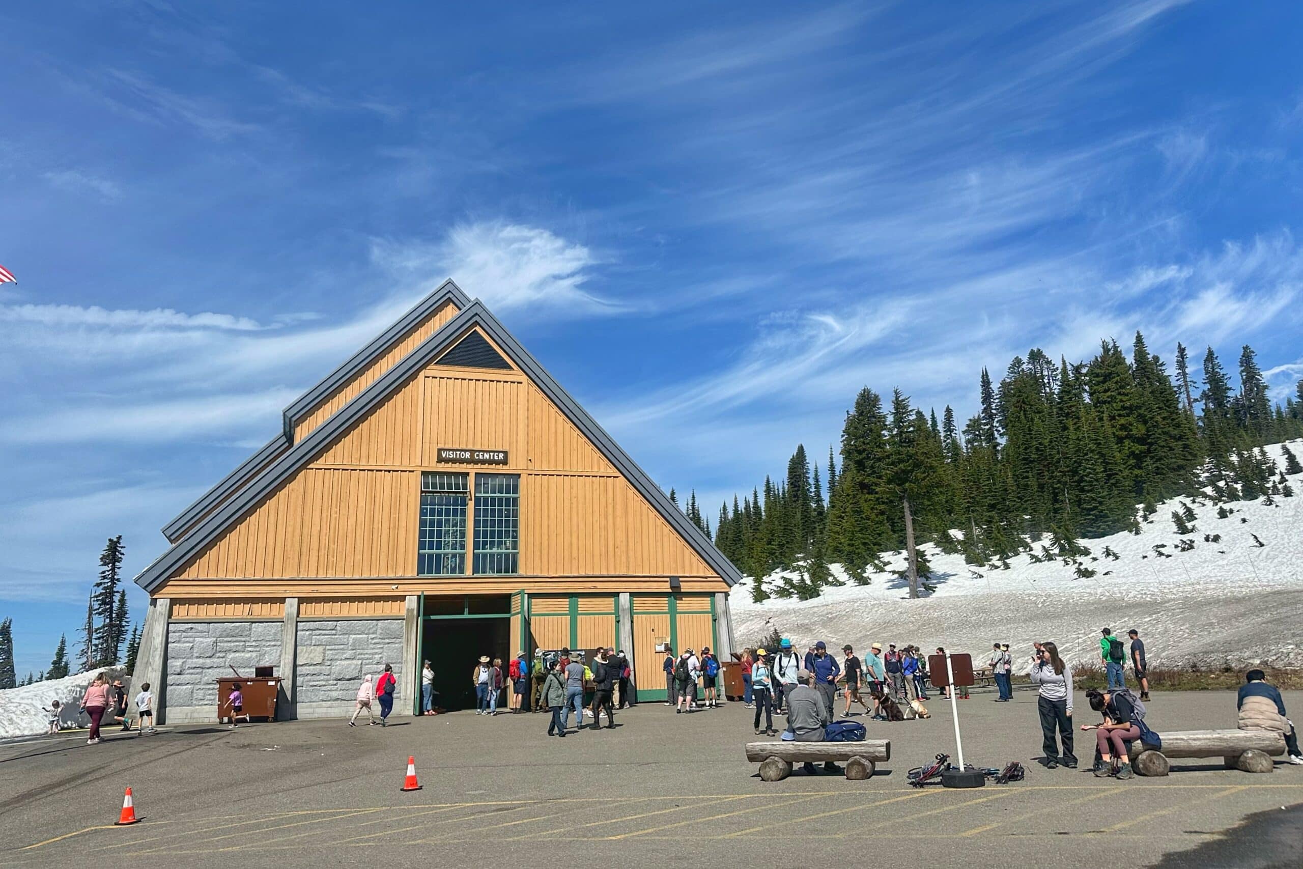 Guests gather outside the visitor center at Mt Rainier in June with deep snow still covering the ground around the parking lot. Tall evergreen trees line the hillside behind the building beneath a bright blue sky streaked with wispy clouds.