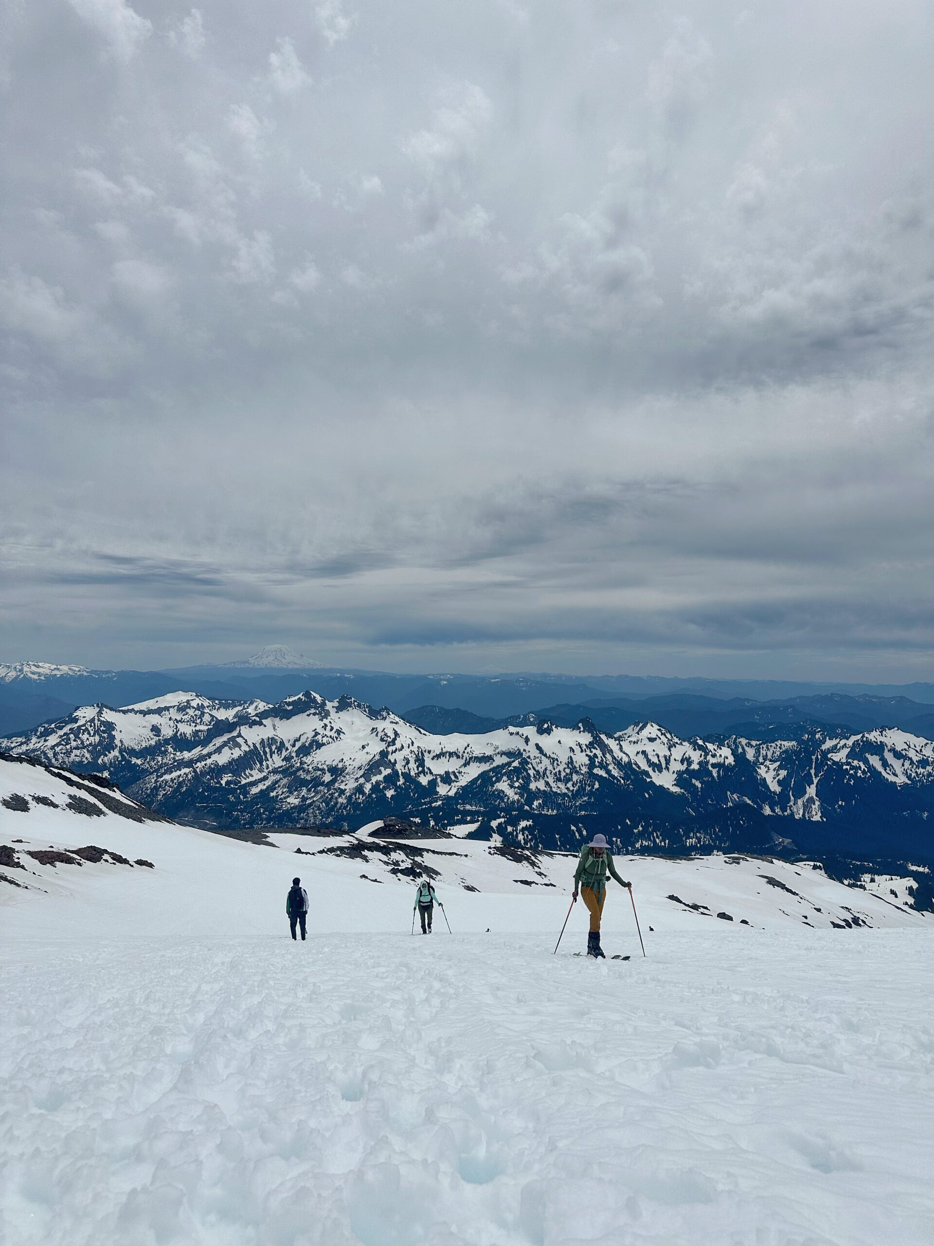 Hikers crossing a snow-covered slope show what Paradise trail conditions in June at Mount Rainier can look like, with rugged mountain peaks stretching into the distance beneath thick gray clouds.