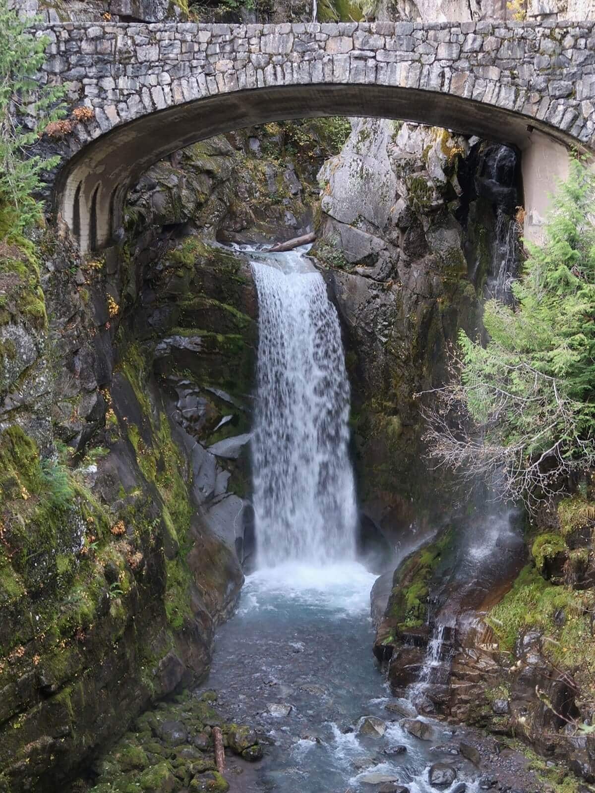 Stone arch bridge spanning Christine Falls as the waterfall drops through a narrow rocky gorge into a clear blue pool below. Moss covered cliffs and evergreen trees frame the scene.