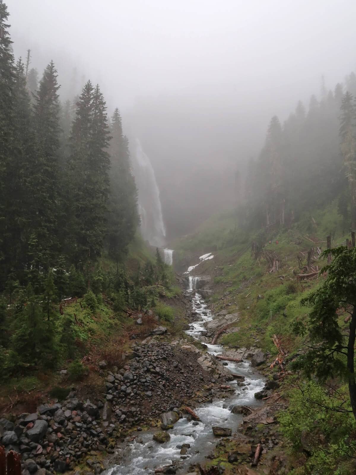 Comet Falls cascades down a steep mountainside through a misty forest, with a rocky creek winding through the green valley below. Low clouds and fog partially veil the waterfall and surrounding evergreen trees, creating a moody view.