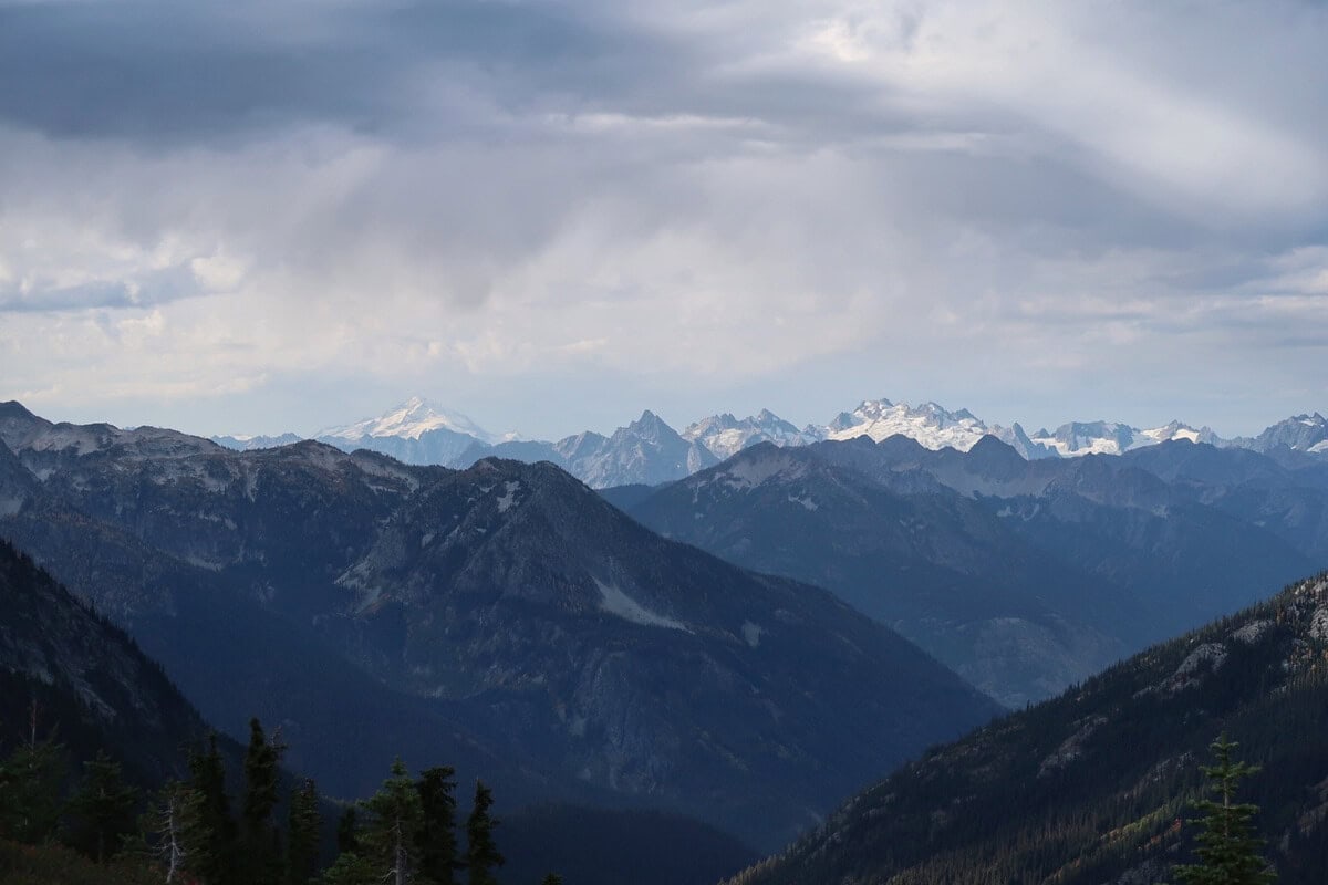 A sweeping view of Glacier Peak from Maple Pass captures layers of rugged mountain ridges beneath dramatic storm clouds, with the snow covered volcano rising on the distant horizon.