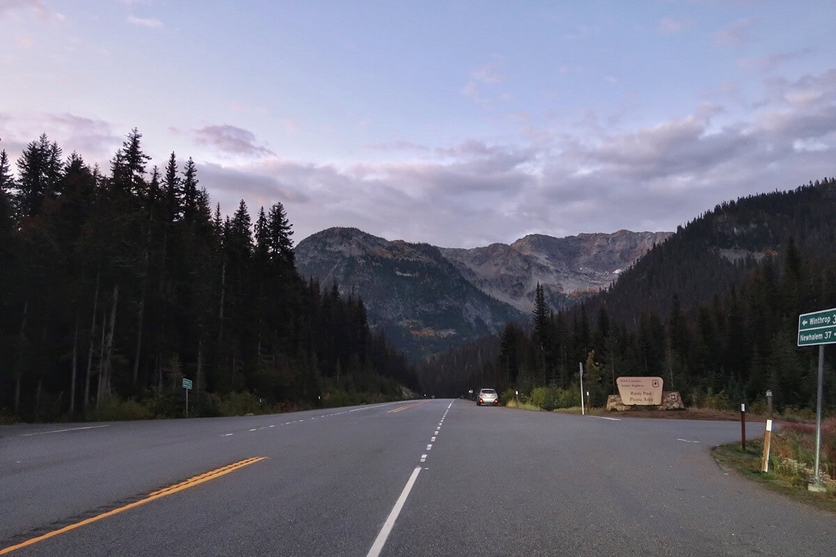 Highway 20 in the North Cascades stretches toward rugged mountain peaks under a soft dawn sky, with evergreen forests lining both sides of the road. A roadside sign marks the Rainy Pass Picnic Area.