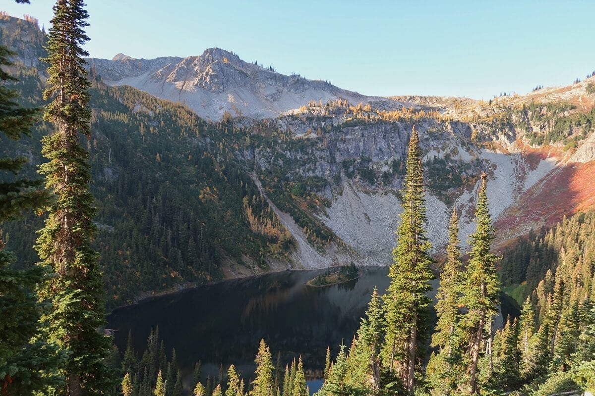 A scenic overlook above Lake Ann in the North Cascades reveals a deep blue alpine lake framed by steep rocky cliffs, evergreen forests, and rugged mountain peaks.