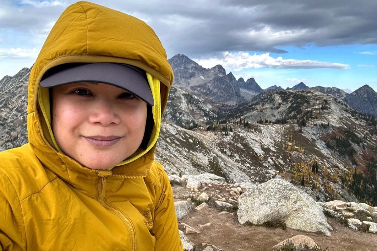 A hiker wearing a yellow hooded jacket and cap pauses for a selfie on the Maple Pass Loop, with rugged granite peaks and rolling alpine ridges stretching into the distance.
