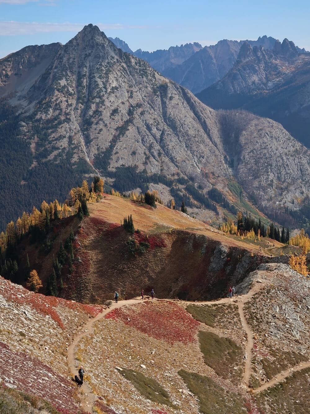 Hikers follow a winding alpine path along the Maple Pass Trail in fall, surrounded by vibrant red and gold vegetation with rugged granite peaks rising in the background.