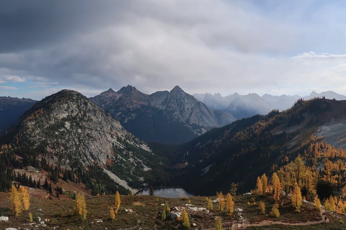 A sweeping view from the Heather - Maple Pass Trail overlooks a small alpine lake surrounded by rugged mountain peaks, golden larch trees, and colorful heather covering the rocky slopes.