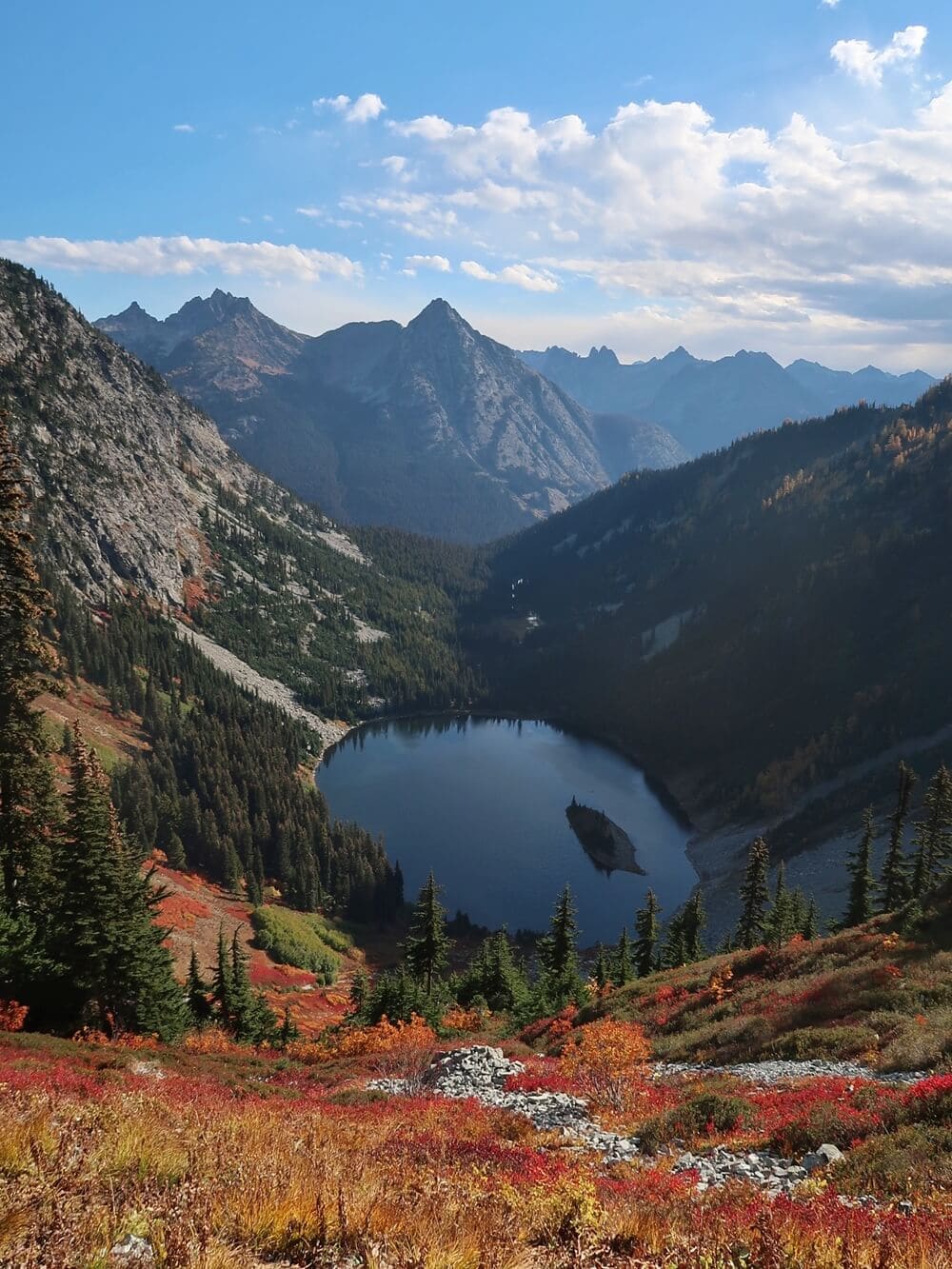 A sweeping view from the Maple Pass Loop Trail overlooks a deep blue alpine lake surrounded by evergreen forests, rugged mountain peaks, and colorful fall foliage.