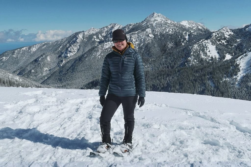 An expansive winter scene from a Hurricane Ridge snowshoe tour featuring a woman wearing winter hiking gear standing on a snowy ridge with rugged mountains and evergreen forests behind her.