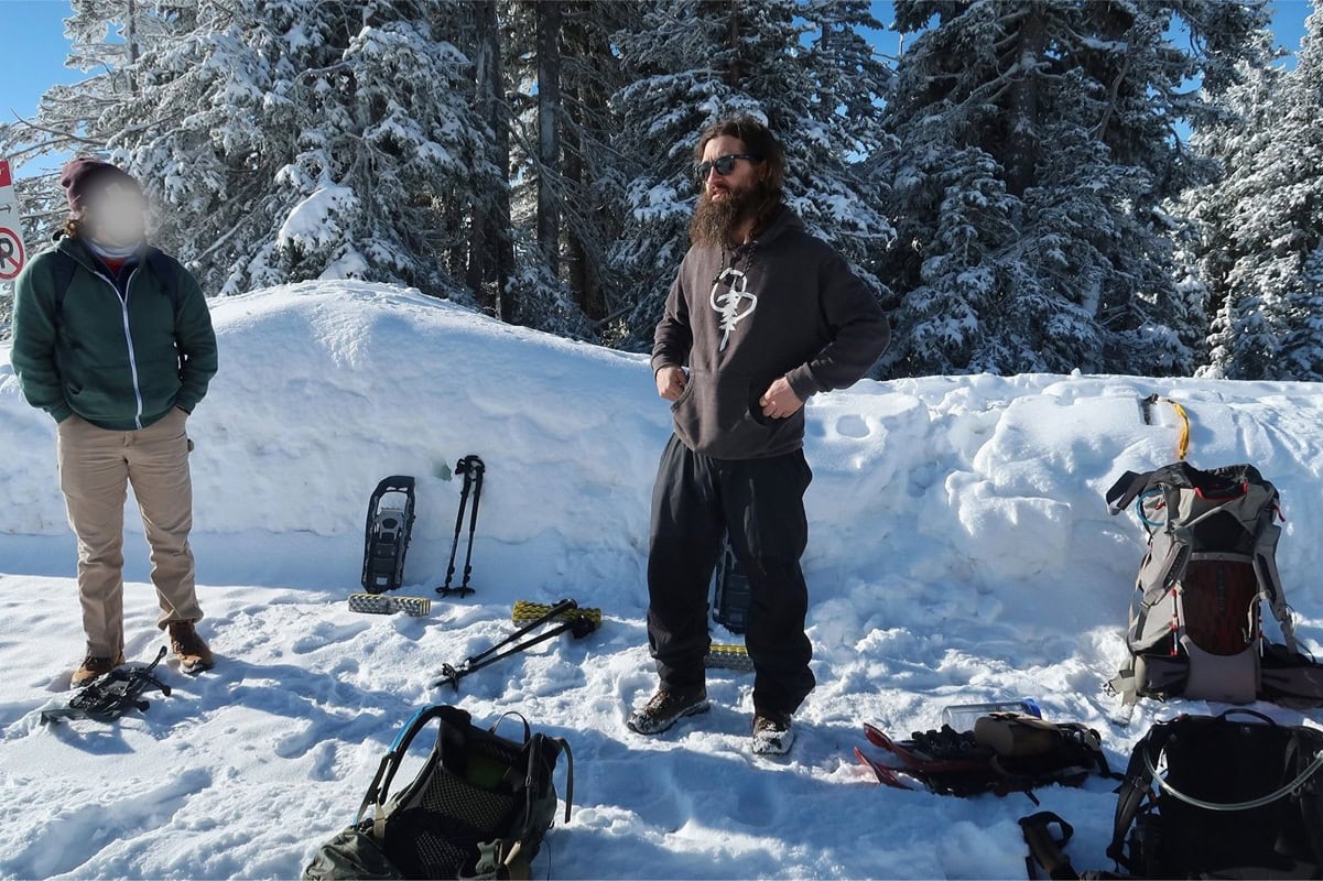Two men stand in deep snow beside snowshoes, trekking poles, and backpacks while preparing for a winter hike. Snow covered evergreen trees surround the group under a clear blue sky.
