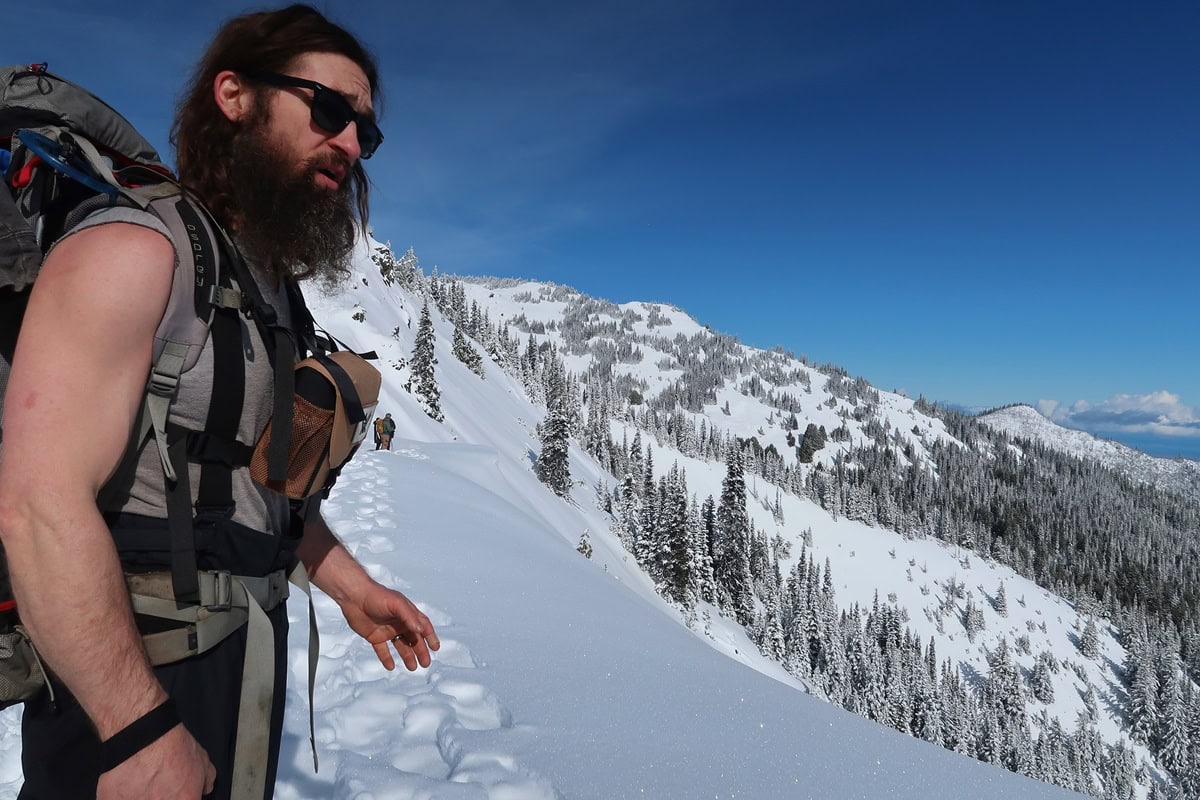 A guide points out a cornice, one winter hazard to be aware of while snowshoeing in Olympic National Park. A steep snow covered mountainside and evergreen forest stretch behind him under a clear blue sky.