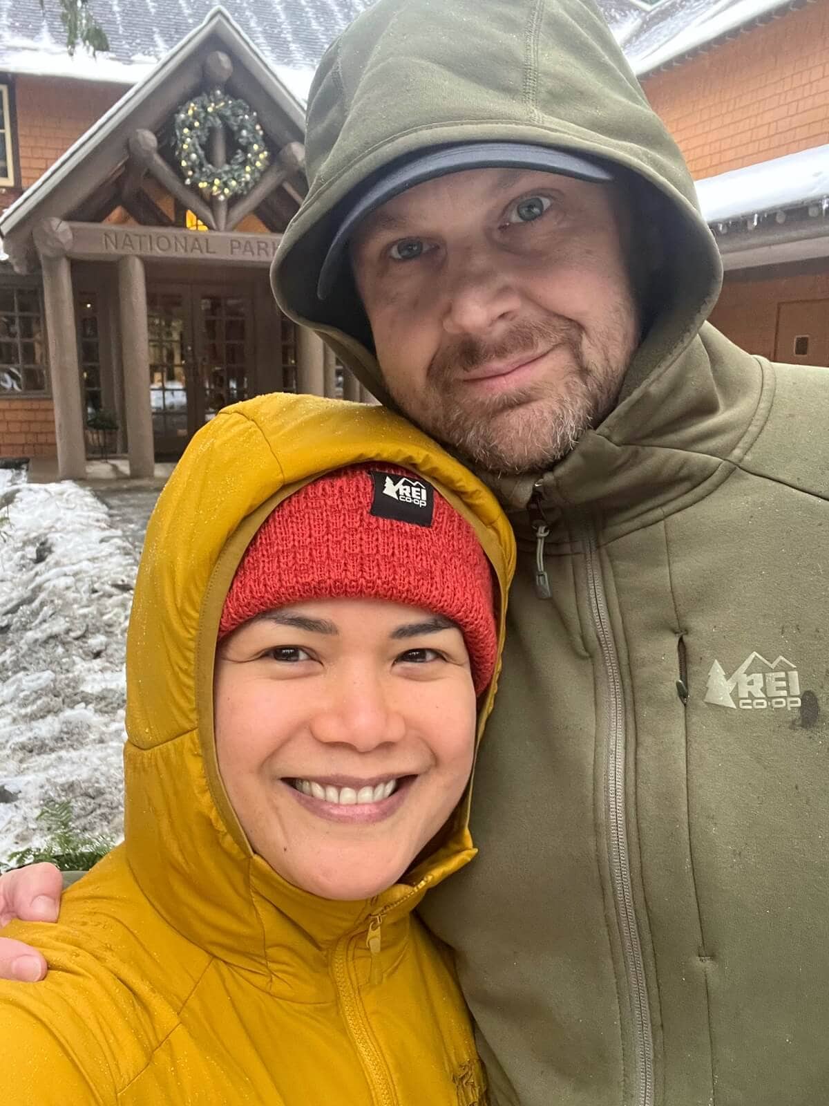 A smiling couple wearing hooded jackets poses for a selfie outside a national park building on a wet and snowy day. The woman wears a yellow Arc’teryx Atom Hoody in light rain, while the man wears an olive green REI Co-op jacket.