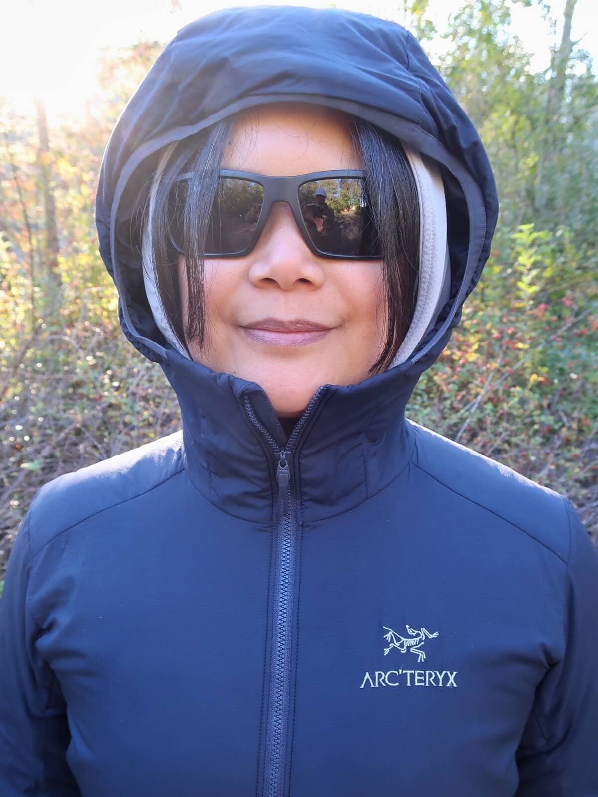 A hiker wearing a navy blue Arc'teryx Atom Hoody zipped up to her chin with the hood pulled over a gray sun hoody. She wears black sunglasses and stands outdoors with autumn foliage in the background.