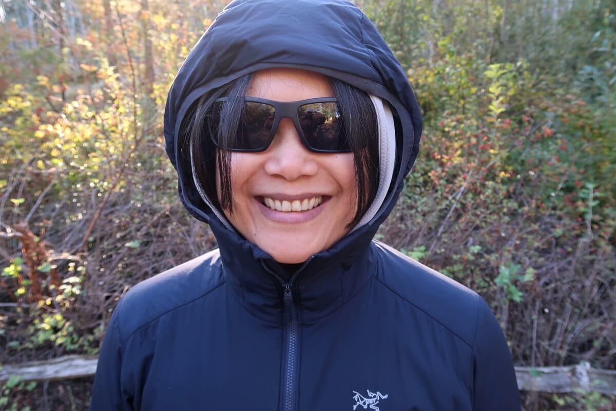 A smiling hiker wearing a navy blue Arc'teryx Atom Hoody zipped up to her chin with the hood pulled over a gray sun hoody. She wears black sunglasses while showing off the fit of the hiking jacket on a wooded trail